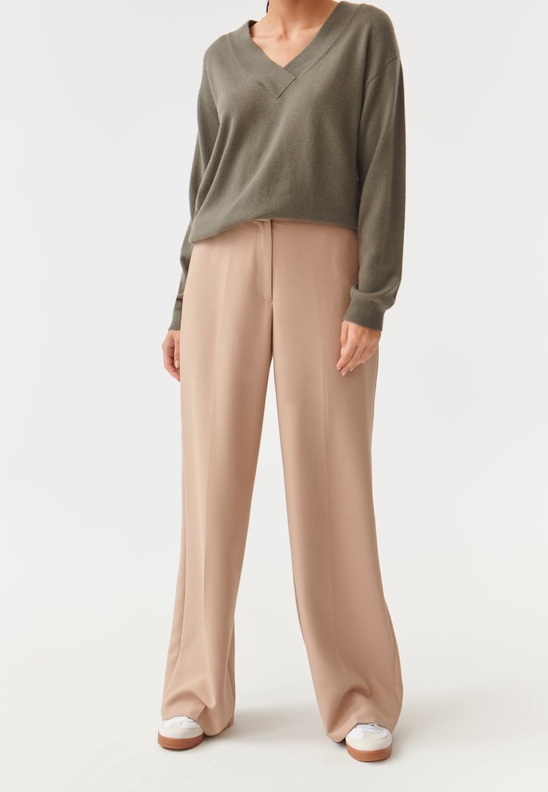 Personne portant un pull col en V vert olive, un pantalon beige à jambes larges et des baskets blanches sur un fond uni.