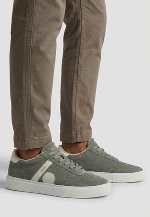 Osoba nosiaca sivé semišové tenisky s bielymi podrážkami a béžovými akcentmi, v kombinácii so svetlohnedými nohavicami.