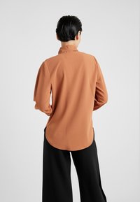 Blouse marron à manches longues avec une texture lisse, col et ourlet arrondi, associée à un pantalon noir à jambes larges.