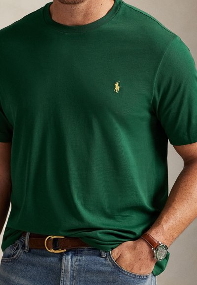 Πράσινο βαμβακερό μπλουζάκι με στρογγυλή λαιμόκοψη και μικρό κίτρινο κεντημένο λογότυπο στο αριστερό στήθος, συνδυασμένο με καφέ ζώνη και ρολόι.