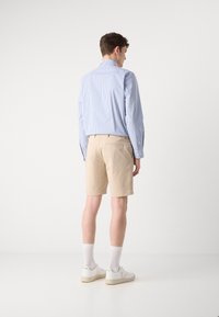 Lys beige shorts med en skreddersydd passform, kombinert med en lys blåstripete langermet skjorte og hvite sneakers, sett fra baksiden.