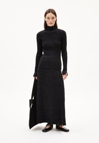 Maglione a collo alto nero a coste abbinato a una lunga gonna coordinata, entrambi con un sottile motivo a macchie. Ballerine nere e una borsa a tracolla completano l'outfit.