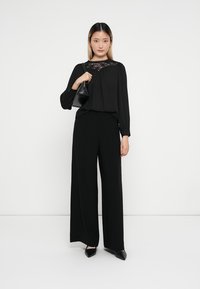 Blusa preta com detalhes em renda na gola, combinada com calças pretas de perna larga. A modelo segura uma pequena mala preta. Os sapatos são de bico fino.