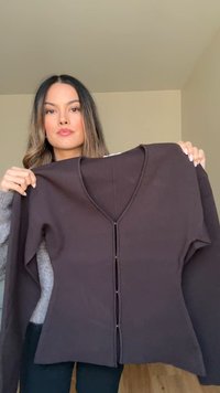 Vrouw met lang haar houdt een donkerbruine V-hals cardigan met knoopsluiting aan de voorkant tegen een effen beige muur.