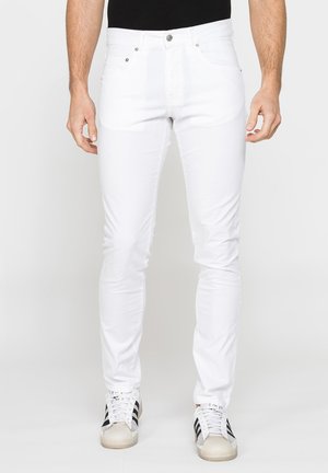 Uomo che indossa jeans bianchi slim fit, top nero e sneakers bianche con strisce nere, in piedi davanti a uno sfondo uniforme.