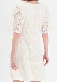 Robe en dentelle avec manches courtes bouffantes, de couleur blanche, présentant des motifs complexes en forme de diamants et de fleurs. Tissu doux et texturé avec une coupe ample.