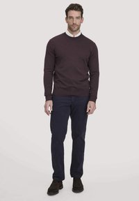 Bordeauxfarbener Rundhalsausschnitt-Pullover mit gerippten Bündchen, kombiniert mit blauen Hosen. Modell steht vor einem neutralen Hintergrund und trägt braune Schuhe.