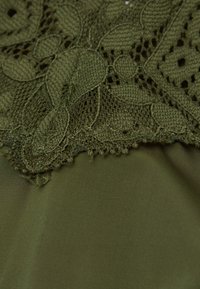 Zielony materiał z misterną koronką, zawierającą wzory liści. Krawędź koronki jest falbaniasta, kontrastując z gładkim, podłożonym materiałem.