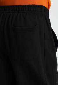 Pantalones de chándal negros con una cintura elástica, que cuentan con un bolsillo trasero y una textura suave, parcialmente mostrados junto a una camiseta naranja.