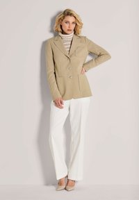 Blonde vrouw in een beige blazer, gestreepte coltrui, witte broek en beige hakken, staande met haar hand op de heup tegen een lichte achtergrond.
