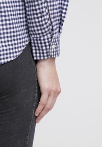 Camicia a quadri blu e bianchi con polsini abbottonati, abbinata a jeans grigi scuri. Il tessuto ha una consistenza liscia e un motivo classico.