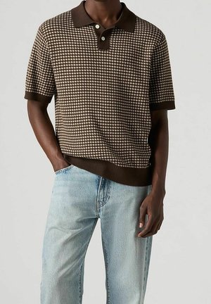 Homme portant un polo à manches courtes marron et beige à motifs, associé à un jean bleu clair, debout avec une main dans la poche.
