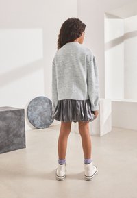 Maglione lavorato a maglia grigio chiaro, gonna plissettata grigio scuro, scarpe da ginnastica bianche con strisce nere e calzini viola. Vista da dietro.