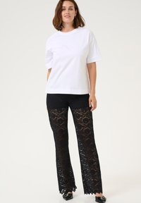Witte katoenen t-shirt met korte mouwen, gecombineerd met zwarte lingerieachtige broek met bloemenpatronen en gegolfde randen, gedragen met zwarte schoenen.