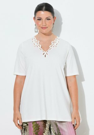 Kvinde med mørkt hår iført en hvid top med korte ærmer og udskæring ved halsudskæringen, iført mønstrede lyserøde og grønne bukser og store rektangulære øreringe.