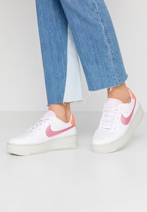 Personne portant des baskets à plateforme blanches avec un swoosh rose et un onglet de talon orange, associées à un jean denim bleu bicolore raccourci.