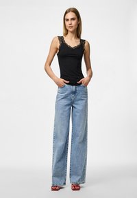 Svart tank top med blondekant, kombinert med lyseblå vide jeans. Modellen står med hendene i lommene, iført røde sandaler.