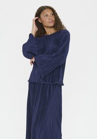 Námornícky dvojdielny outfit so skladanými detailmi a dlhými rukávmi, ozdobený volánikom. Mäkko tkanina elegantne splýva cez telo.