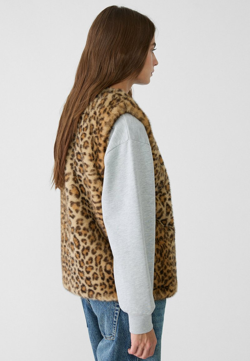 Gilet in pelliccia sintetica con stampa leopardata abbinato a una leggera felpa grigia a maniche lunghe, visto di lato. Sono visibili dei pantaloni di jeans casual.