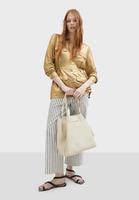 Guldmetallisk skjorta med knappar, randiga vida byxor, svarta sandaler och en texturerad krämfärgad tote-väska.