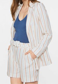 Kvinna iförd vita högmidjade shorts och matchande kavaj med persika- och svarta vertikala ränder, tillsammans med en blå ärmlös topp.