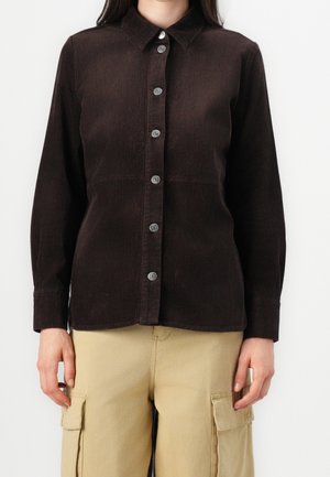 Kvinde iført en mørkebrun kordfløjls-knapbluse og beige cargobukser, stående med armene afslappet ned langs siden.