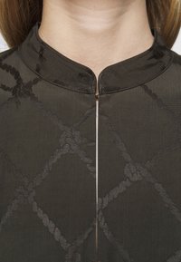 Blusa verde oscuro con cuello alto, que presenta un patrón de cuerda texturizado, cremallera central y tejido suave para un aspecto entallado.