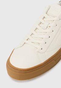 White leather sneaker with rounded toe, flat laces, and rubber gum sole. Features subtle branding on the side and textured detail on the sole.