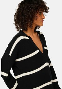 Femme avec des cheveux bouclés portant un pull en tricot noir avec des rayures blanches horizontales et un col en V profond, de profil sur un fond blanc.