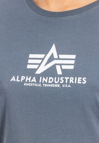 Siva bombažna majica s krožno izrezom. Ponuja bel logo z oglatim zvezdo in črtami, besedilo pa se glasi "ALPHA INDUSTRIES" in podrobnosti o lokaciji.