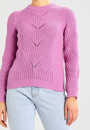 Femme portant un pull en maille texturée violet clair avec un motif chevron subtil, associé à un jean bleu clair sur un fond neutre.