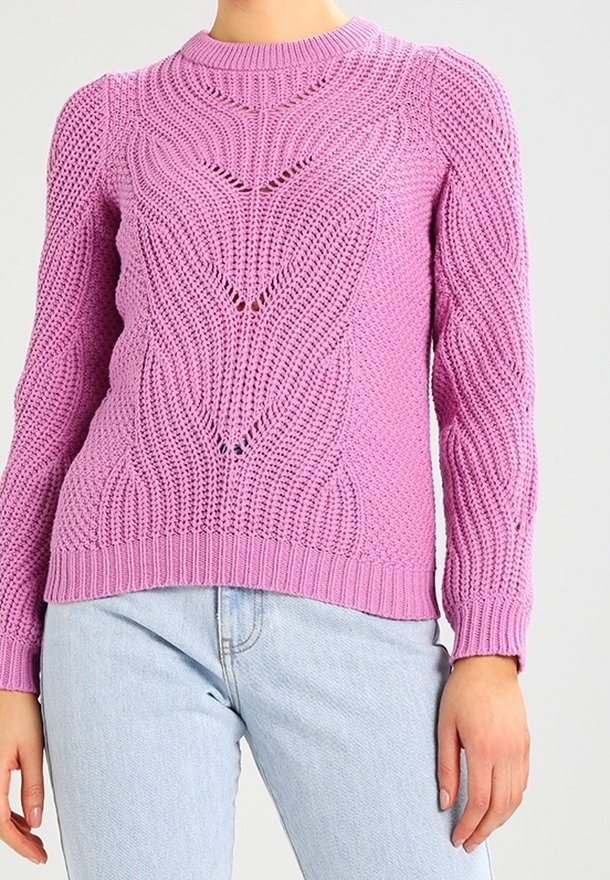 Femme portant un pull en maille texturée violet clair avec un motif chevron subtil, associé à un jean bleu clair sur un fond neutre.