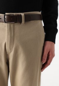 Beige Baumwollhose mit geradem Schnitt. Kombiniert mit einem dunkelbraunen Ledergürtel mit einer silbernen Schnalle. Glatte Textur.