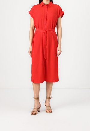 Kobieta ubrana w czerwoną koszulową sukienkę zapinaną na guziki, sięgającą do kolan, z paskiem w talii oraz beżowe sandały na wysokim obcasie z paskami.