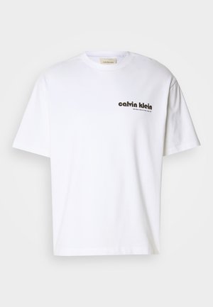 White cotton t-shirt with short sleeves, featuring black "Calvin Klein" text logo and a square tag near the neckline. Simple design.