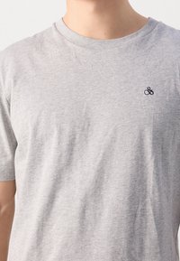 Grijze katoenen T-shirt met een ronde hals. Heeft korte mouwen en een klein zwart geborduurd logo op de linkerkant van de borst.