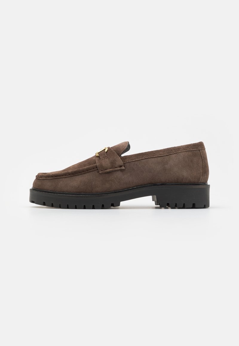 Mocassin en daim marron avec une semelle en caoutchouc noir texturé et un détail de boucle dorée décoratif à l'avant.