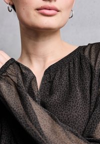 Schwarze Bluse aus durchsichtigem Stoff mit Leopardenmuster. Verfügt über einen V-Ausschnitt, geraffte Details und lange Ärmel mit elastischen Bündchen. Mit Reißverschluss vorne.