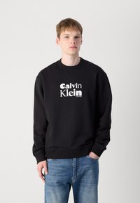 Black cotton sweatshirt featuring a white "Calvin Klein" logo in bold typography. Crew neck and ribbed cuffs with a relaxed fit.