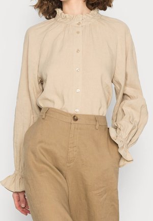 Femme portant un chemisier beige à col à volants avec manches bouffantes et un pantalon taille haute couleur fauve avec passants de ceinture et fermeture à bouton sur le devant.