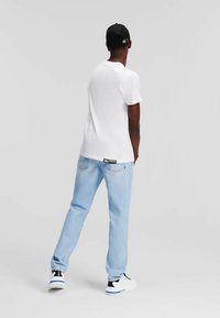 Camiseta blanca de algodón, jeans de mezclilla azul claro y zapatillas negras con acentos azules, vistas desde atrás contra un fondo gris claro.