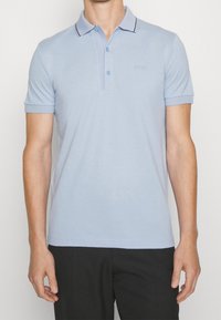 Muž v světle modrém polo tričku s krátkým rukávem, knoflíkovým límcem a logem "BOSS" na hrudi, v kombinaci s černými kalhotami.