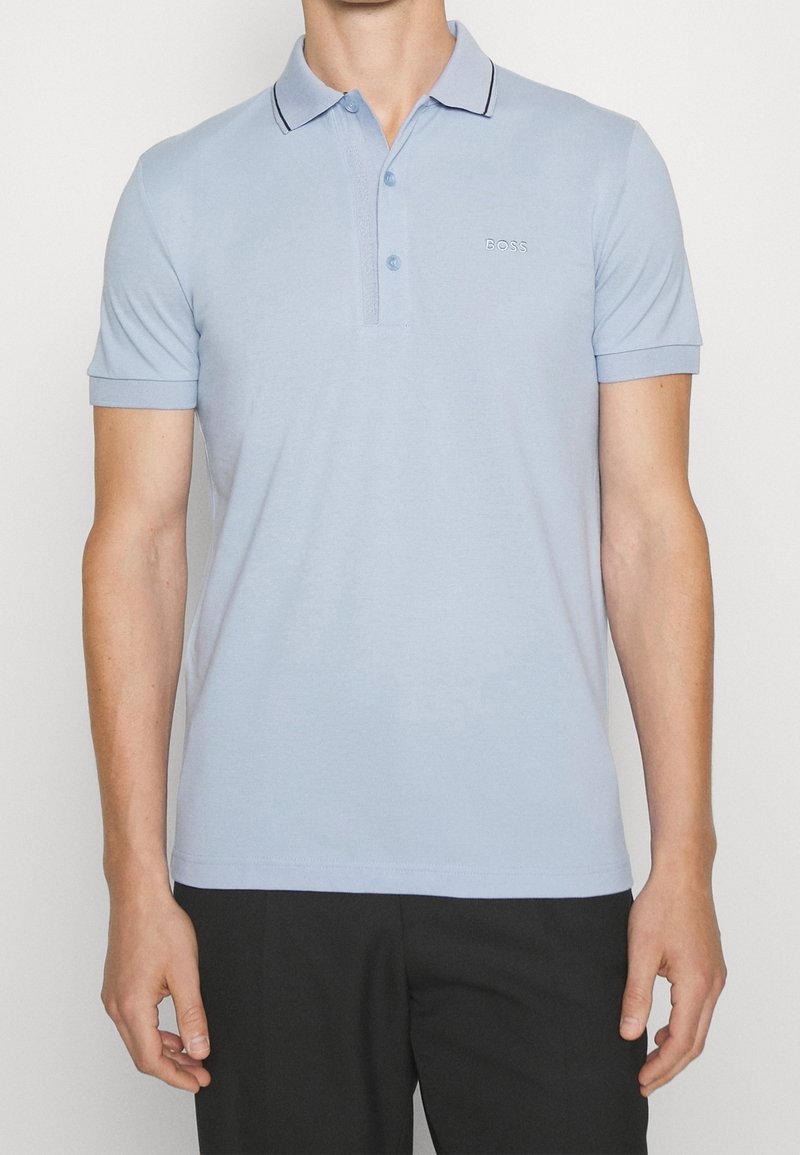 Muž v světle modrém polo tričku s krátkým rukávem, knoflíkovým límcem a logem "BOSS" na hrudi, v kombinaci s černými kalhotami.