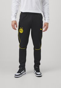 Man in zwarte Puma trainingsbroek met geel Dortmund BVB 09-logo en gele piping, gecombineerd met zwart-witte sneakers.