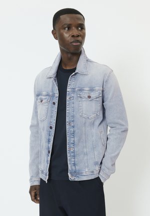 Ung mand iført lys blå denimjakke og sort skjorte, stående med den ene hånd i lommen, kiggende til siden mod en hvid baggrund.