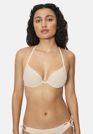 Mujer con cabello rizado y oscuro, vistiendo un bikini crema texturizado con top halter y bottom con lazos laterales, sobre un fondo blanco liso.