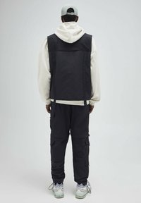 Czarna kamizelka na białej bluzie z kapturem, w połączeniu z czarnymi spodniami typu cargo. Jasnoszara czapka. Widoczne detale to siateczkowe akcenty i liczne kieszenie.