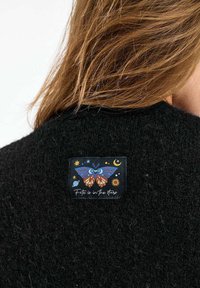 Osoba so svetlohnedými vlasmi oblečená v čiernom huňatom odeve s nášivkou zobrazujúcou farebného motýľa a hviezdy, text znie „Osud je v hviezdach.“