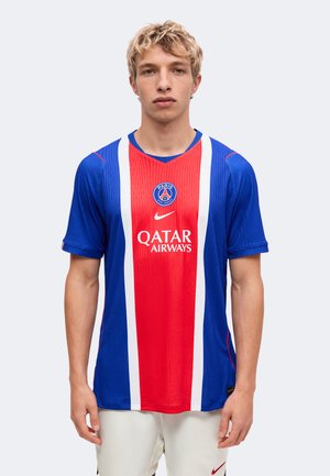 Jeune homme portant un maillot de football bleu et rouge du Paris Saint-Germain avec le logo Qatar Airways, debout sur un fond clair uni.
