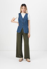 Jeansvest med v-hals og knaplukning, båret over en hvid t-shirt, parret med brede olivengrønne bukser og sko i afslappet stil.
